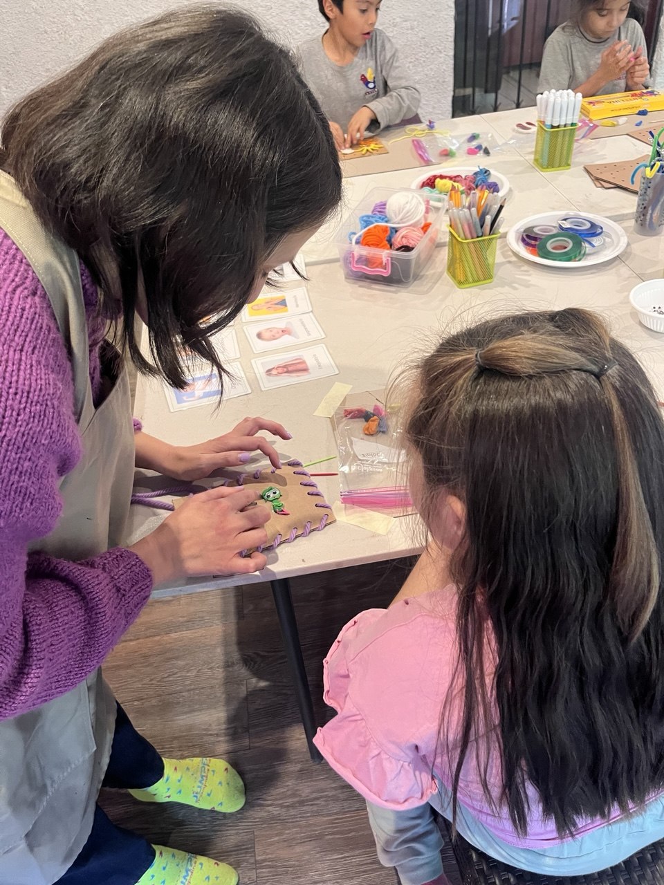 Tallerista ayudando a una niña a tejer con lana sobre cartón